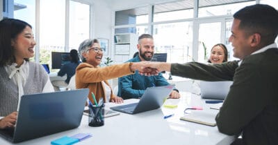 Fünf Personen in einem modernen Büro, die an einem Tisch sitzen und sich die Hände schütteln.